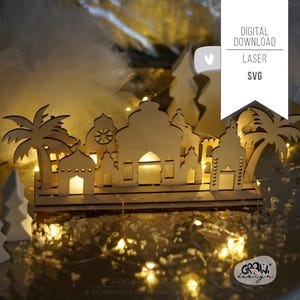 May include: A laser cut wooden silhouette of a Middle Eastern city with palm trees and a star on top of the tallest building. The silhouette is lit up with fairy lights and is sitting on a white surface. The text "DIGITAL DOWNLOAD LASER SVG" is in the top right corner of the image.
