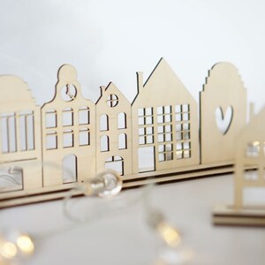 May include: A wooden cutout of a row of houses with a heart cutout on one of the houses. The houses are arranged on a wooden base and are illuminated with string lights.
