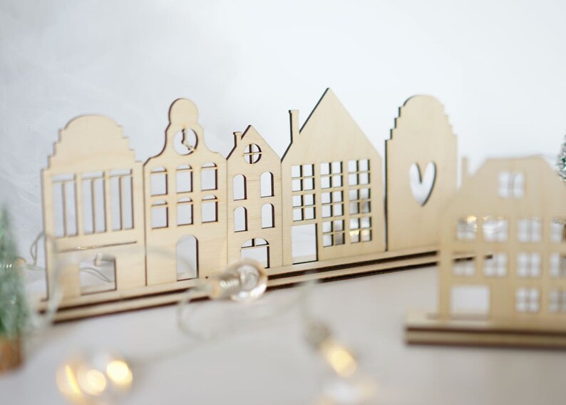May include: A wooden cutout of a row of houses with a heart cutout on one of the houses. The houses are arranged on a wooden base and are illuminated with string lights.