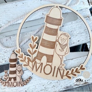 Może przedstawiać: Drewniana dekoracja ścienna w stylu marynistycznym z latarnią morską, ptakiem i napisem "Moin" w kółku. Pokazano również mniejszą wersję dekoracji.