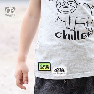 K&ouml;nnte beinhalten: Ein graues T-Shirt mit einer schwarz-wei&szlig;en Illustration eines Faultiers und dem Text "chiller". Das Shirt hat einen gr&uuml;n-gelben Patch mit der Aufschrift "l&auml;ssig" und einen wei&szlig;en Patch mit der Aufschrift "grow design".