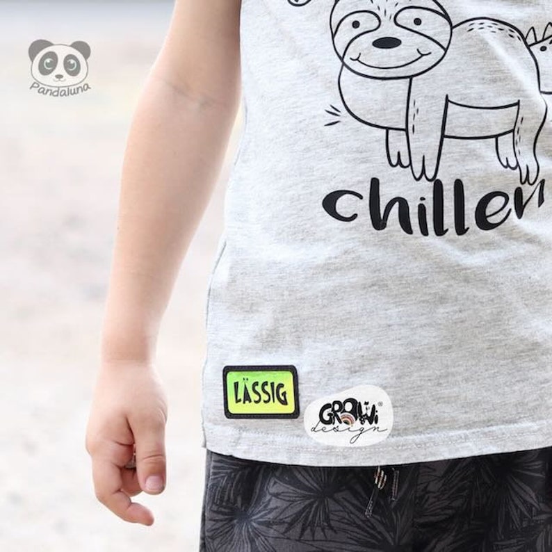 K&ouml;nnte beinhalten: Ein graues T-Shirt mit einer schwarz-wei&szlig;en Illustration eines Faultiers und dem Text "chiller". Das Shirt hat einen gr&uuml;n-gelben Patch mit der Aufschrift "l&auml;ssig" und einen wei&szlig;en Patch mit der Aufschrift "grow design".