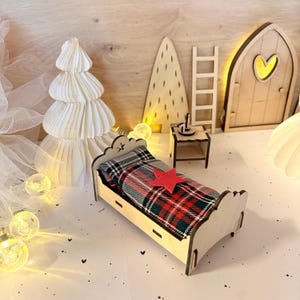 May include: A miniature wooden bed with a plaid blanket and a red star. The bed is part of a Christmas-themed display, which includes a paper tree, a wooden door with a heart-shaped window, and fairy lights.