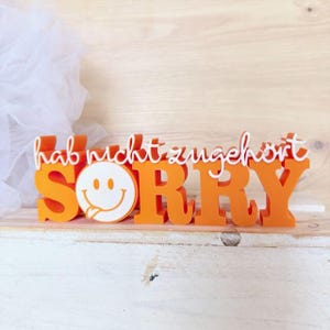 May include: Orange wooden sign with the word "SORRY" in large capital letters. The letter "O" is a white smiley face with its tongue sticking out. The words "hab nicht zugehört" are written above the word "SORRY".