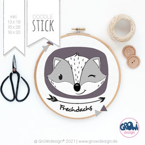 May include: A purple and gray embroidery hoop with a cartoon fox face and the text "Frechdachs" in black. The fox is winking and has a happy expression. The hoop is surrounded by white triangles.