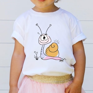 May include: White t-shirt featuring a cartoon snail design. The snail has a light pink body, a tan shell, and a smiling face with heart-shaped antennae. The design includes small hearts and blades of grass.
