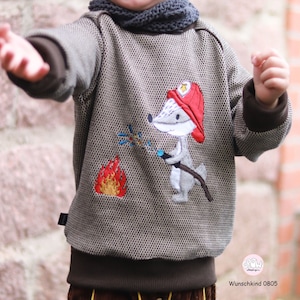 Könnte beinhalten: Ein brauner Pullover mit einem braunen und weißen Fuchs, der einen roten Feuerwehrhelm trägt und einen Schlauch hält. Der Fuchs steht vor einer gelben und orangen Flamme. Der Pullover hat den Text "Wunschkind 0805" unten.