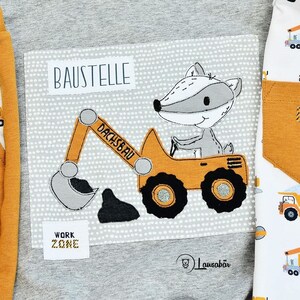 May include: A gray and white fabric patch with a yellow excavator and a gray badger driving it. The text "BAUSTELLE" is above the excavator and "WORK ZONE" is below. The text "DACHSBau" is on the excavator's arm.