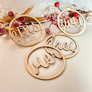 May include: Four wooden Christmas ornaments with the numbers one through four written in German on them. The ornaments are round and have a hole at the top for hanging.