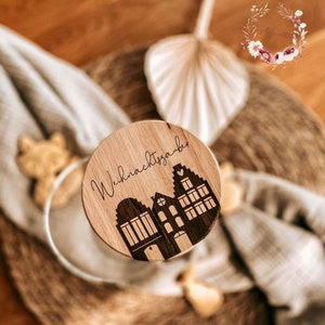 May include: A round wooden ornament with the text "Weihnachtszauber" and a silhouette of three houses with hearts on the windows.
