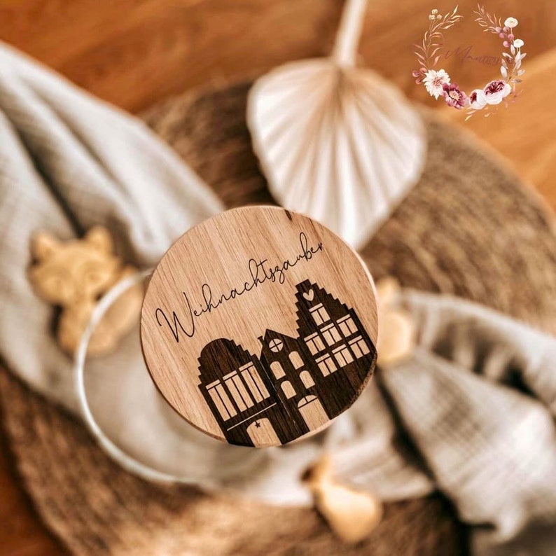 May include: A round wooden ornament with the text "Weihnachtszauber" and a silhouette of three houses with hearts on the windows.