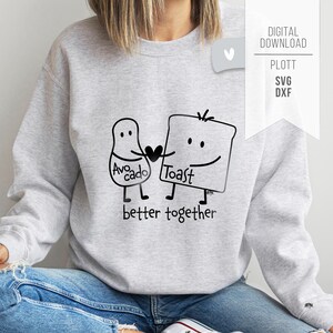 以下が含まれることがあります： アボカドとトーストがハートを持って手をつないでいる白黒のイラストが描かれた、明るい灰色のスウェットシャツ。「Avocado」と「Toast」の文字がキャラクターに書かれており、「better together」というフレーズが下にあります。「DIGITAL DOWNLOAD」と「PLOTT SVG DXF」の文字が白いタグに書かれています。