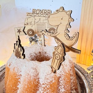 May include: Wooden cake topper set featuring a dinosaur in a fire truck with the word "ROARR" above it. The set also includes a fire hydrant and a flame.