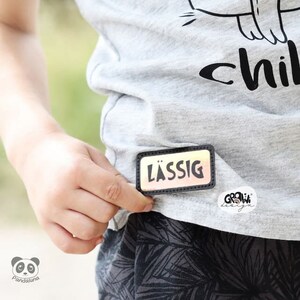 K&ouml;nnte beinhalten: Ein graues T-Shirt mit schwarzem Text, der "child" sagt, und einer schwarz-wei&szlig;en Grafik einer Katze. Das Shirt hat einen schwarzen und orangen Patch, auf dem "l&auml;ssig" steht.
