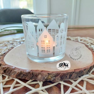 May include: A white glass candle holder with a cityscape design. The candle holder is sitting on a wooden slice with the text "GROW! design" and "ANIRPSI & KNORZA" printed on it.