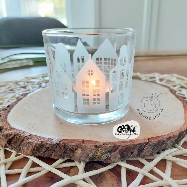 May include: A white glass candle holder with a cityscape design. The candle holder is sitting on a wooden slice with the text "GROW! design" and "ANIRPSI & KNORZA" printed on it.