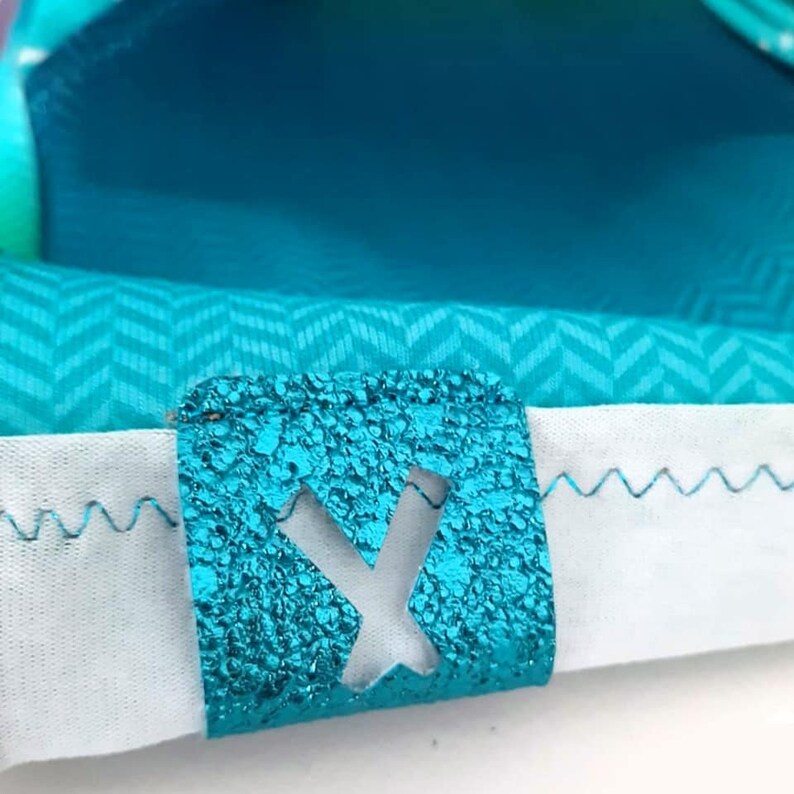 K&ouml;nnte beinhalten: Nahaufnahme eines t&uuml;rkisfarbenen Stoffes mit Chevronmuster. Ein t&uuml;rkisfarbener Stoff-Patch mit einem ausgeschnittenen Buchstaben "Y" ist auf den Stoff gen&auml;ht.