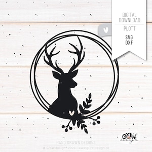 Könnte beinhalten: Eine schwarze Silhouette eines Hirsches mit Geweih in einem Doppelkreis-Kranz mit Blättern und Beeren. Das Design ist auf einem weißen Holzmaserungshintergrund.