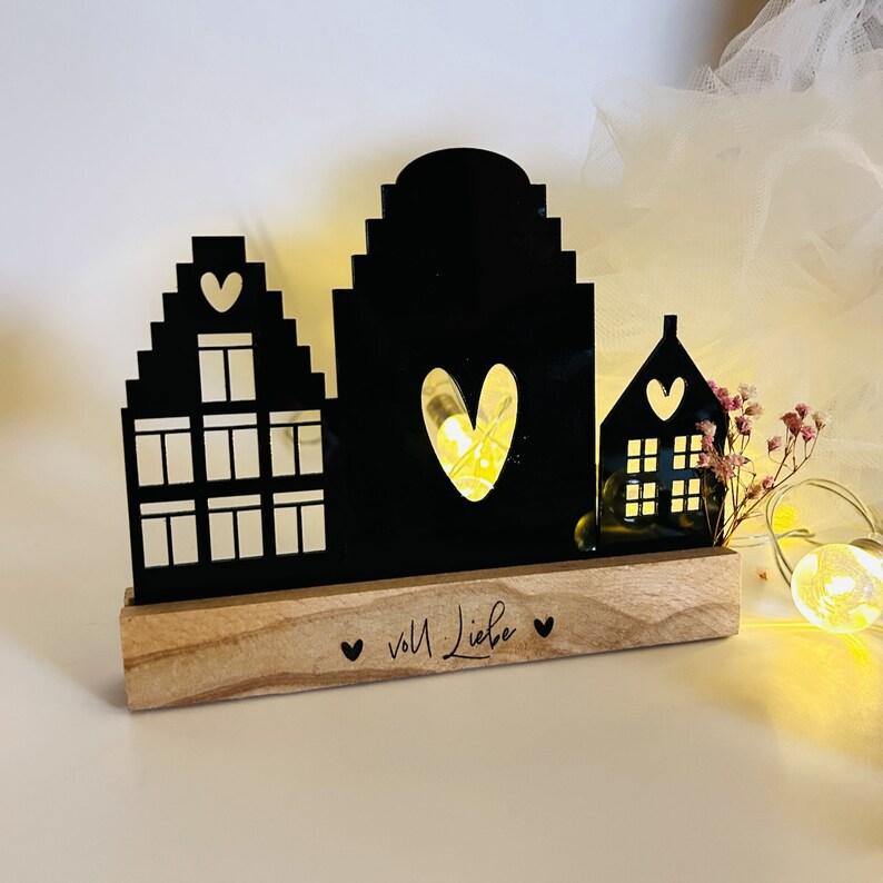 May include: Black silhouette of three houses with a heart cut out of the center house. The houses are on a wooden base with the text "voll Liebe" written on it.
