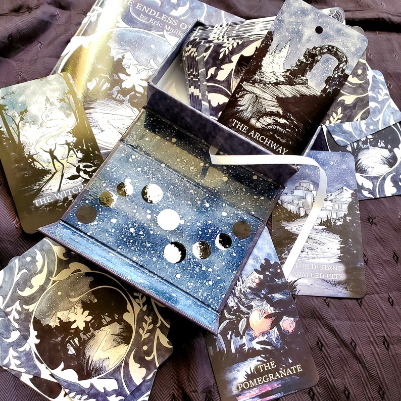 May include: A deck of tarot cards with a celestial theme, featuring illustrations in shades of blue, white, and black. The cards are displayed with their box, which has a starry design and moon phase cutouts. The card titles include "The Witch" and "The Archway."