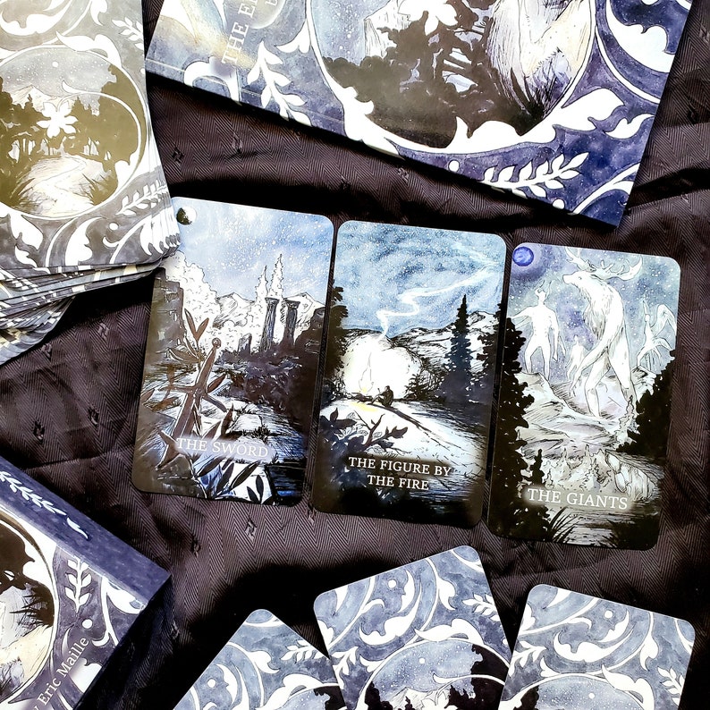 May include: A tarot deck with three cards visible, each featuring a different illustration with text. The cards are on a dark surface, with other cards and a box visible. The text on the cards reads "THE SWORD", "THE FIGURE BY THE FIRE", and "THE GIANTS".