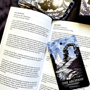 May include: A tarot card with a watercolor illustration of an archway under a starry sky, with the words "THE ARCHWAY" at the bottom. The card is on top of an open book.