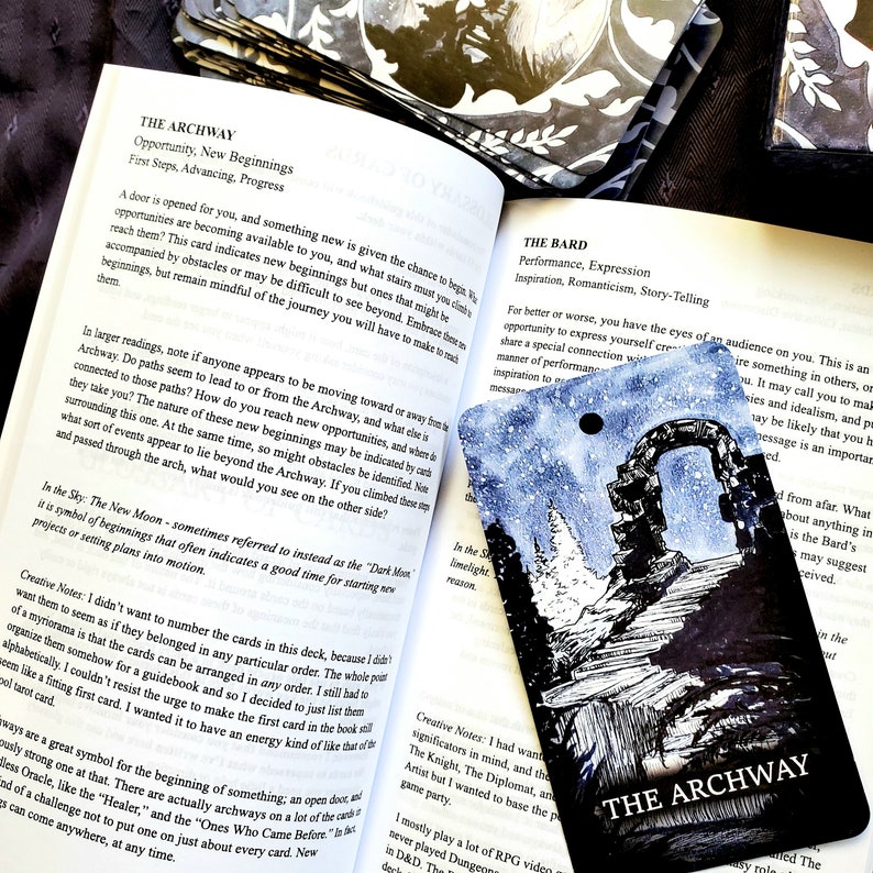 May include: A tarot card with a watercolor illustration of an archway under a starry sky, with the words "THE ARCHWAY" at the bottom. The card is on top of an open book.