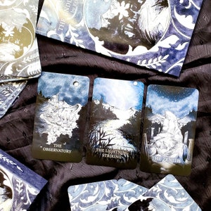 May include: A deck of tarot cards with a celestial theme. Three cards are visible, each featuring a different landscape scene with text: "The Observatory", "The Lightning Striking", and "The Zenith". The cards have a blue and white color scheme, with a starry night sky.