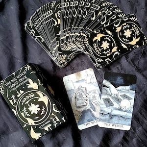 Pu&ograve; includere: Scatola del mazzo di tarocchi The Ink Witch, seconda edizione, con design floreale nero e oro. La scatola &egrave; aperta e mostra le carte a ventaglio. Sono visibili due carte, una con un'illustrazione in bianco e nero di una mano che tiene una carta dei tarocchi e l'altra con un'illustrazione in bianco e nero di una strega con una sfera di cristallo.