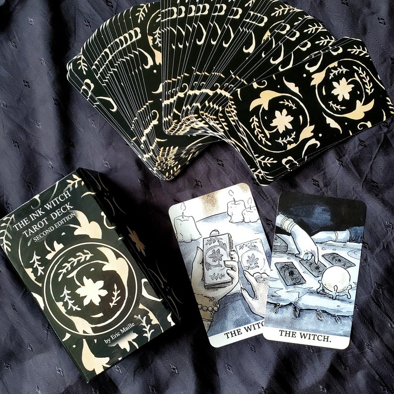 Pu&ograve; includere: Scatola del mazzo di tarocchi The Ink Witch, seconda edizione, con design floreale nero e oro. La scatola &egrave; aperta e mostra le carte a ventaglio. Sono visibili due carte, una con un'illustrazione in bianco e nero di una mano che tiene una carta dei tarocchi e l'altra con un'illustrazione in bianco e nero di una strega con una sfera di cristallo.