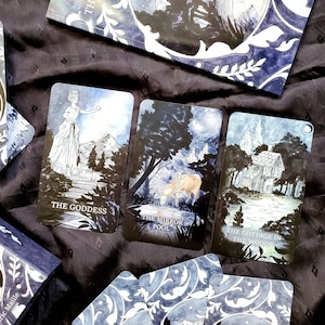 May include: A tarot deck with three illustrated cards featuring the text "THE GODDESS", "THE MIRROR POOL", and "THE COTTAGE IN THE WOODS". The cards have a dark blue and white color scheme with nature-themed illustrations. A book with a matching design is also visible.
