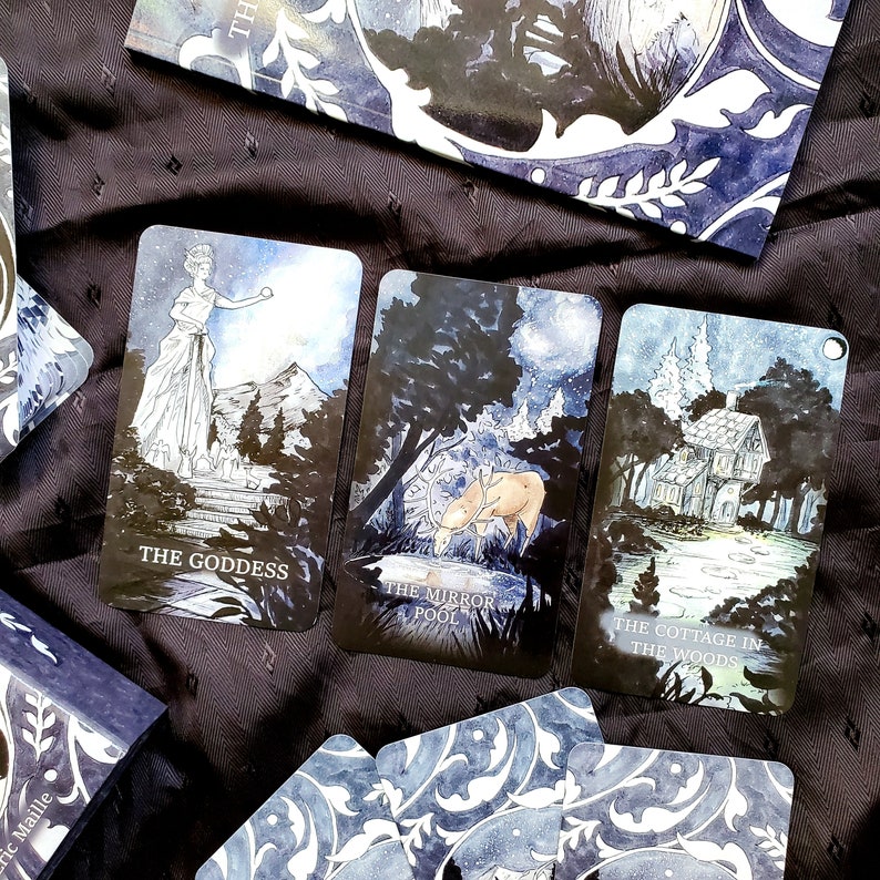 May include: A tarot deck with three illustrated cards featuring the text "THE GODDESS", "THE MIRROR POOL", and "THE COTTAGE IN THE WOODS". The cards have a dark blue and white color scheme with nature-themed illustrations. A book with a matching design is also visible.