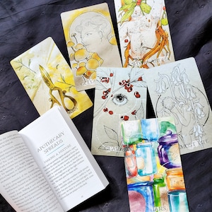 K&ouml;nnte beinhalten: Eine Sammlung von Tarotkarten mit Illustrationen von Blumen, Beeren und einer Person. Die Karten liegen auf einer dunkelgrauen Stoffoberfl&auml;che. Der Text auf den Karten lautet "Gold", "Ashwagandha", "Most Ripe", "Guara" und "Jars".