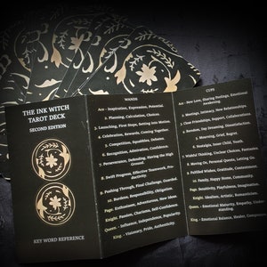Pu&ograve; includere: Un mazzo di tarocchi nero e oro con il titolo "The Ink Witch Tarot Deck, Second Edition" e un riferimento alle parole chiave. Le carte sono disposte a ventaglio, rivelando i nomi dei semi e i significati delle carte per Bastoni e Coppe.