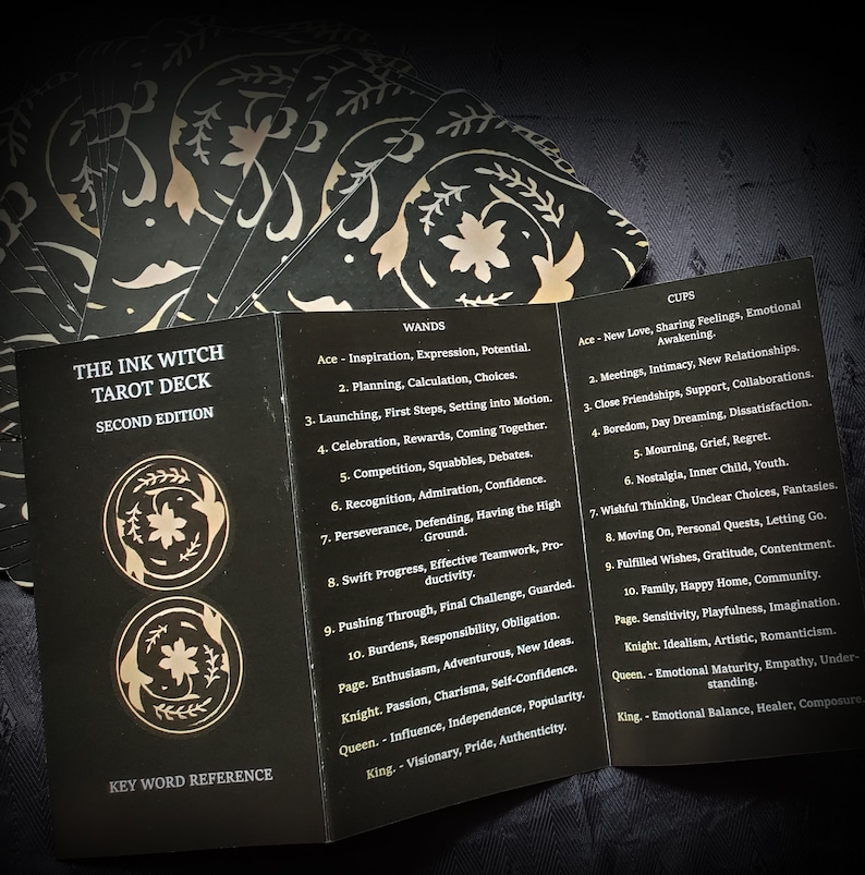 Pu&ograve; includere: Un mazzo di tarocchi nero e oro con il titolo "The Ink Witch Tarot Deck, Second Edition" e un riferimento alle parole chiave. Le carte sono disposte a ventaglio, rivelando i nomi dei semi e i significati delle carte per Bastoni e Coppe.