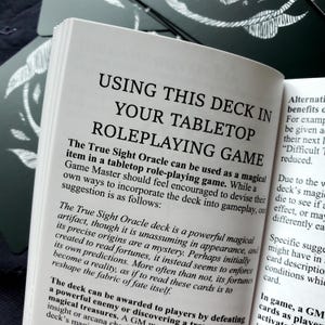 Puede incluir: Libro abierto con el t&iacute;tulo "USING THIS DECK IN YOUR TABLETOP ROLEPLAYING GAME". El texto explica c&oacute;mo usar la baraja True Sight Oracle en un juego de rol de mesa. Se ven otras cartas alrededor del libro.