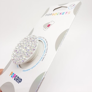 Puede incluir: Agarre para teléfono PopSocket blanco con un diseño de cristal transparente iridiscente. El embalaje dice "PopSockets" y "Top Try Me! Pull Twice to Grip".