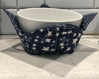 May include: A blue fabric bowl cozy with white stars and a white bowl inside. The cozy is sitting on a white wooden surface.
