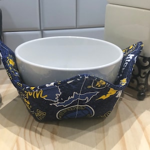 May include: A blue and yellow fabric bowl cozy with a white ceramic bowl inside. The cozy has a pattern of a map and the words "Michigan" and "Go Blue".