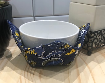 May include: A blue and yellow fabric bowl cozy with a white ceramic bowl inside. The cozy has a pattern of a map and the words "Michigan" and "Go Blue".