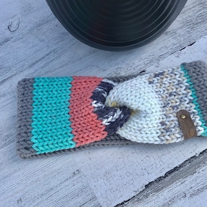 May include: A gray, teal, coral, and white striped knitted headband with a brown leather accent. The headband is knotted in the center.