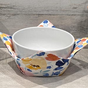 May include: A white ceramic bowl with a floral print bowl cozy. The cozy has a white background with pink, blue, yellow, and orange flowers.