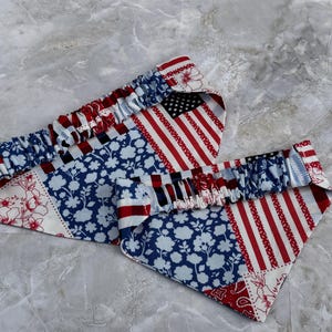 May include: Two triangular fabric pet bandanas featuring a patriotic patchwork design. The pattern includes red, white, and blue sections with floral motifs, stripes, and stars. Each bandana has a gathered elastic neckband for a secure fit.