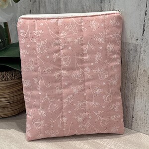 May include: A pink quilted pouch with a white floral pattern and a white zipper. The pouch is rectangular and appears to be made of fabric. The floral design includes various flower and leaf shapes.