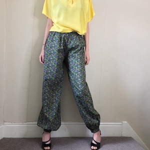 May include: Black pants with a floral print in shades of purple, green, and yellow. The pants have an elastic waistband and a cuffed hem.