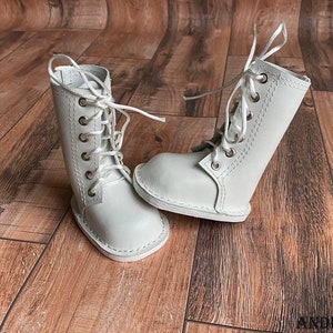 May include: A pair of white leather doll boots with laces. The boots are on a wooden surface.