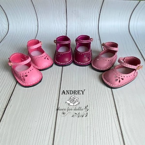 May include: Four pairs of pink and burgundy doll shoes with cut-out designs. The shoes have ankle straps and are made of leather. The shoes are arranged in two rows of two. The shoes are on a light wood surface. The text "ANDREY shoes for dolls" is visible in the image.
