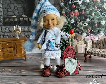 Può includere: Una bambola vestita con un abito invernale, tra cui un cappello a righe blu e bianco con un fiocco di neve, una sciarpa con un disegno di orso polare e stivali marroni. La bambola tiene in mano un piccolo ornamento davanti a un albero di Natale.