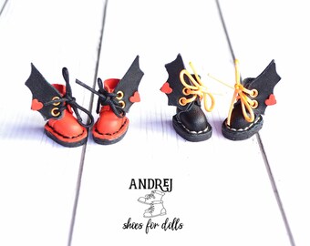 Könnte beinhalten: Zwei Paar Miniatur-Stiefel aus schwarzem Leder mit roten und orangefarbenen Schnürsenkeln. Jeder Stiefel hat einen schwarzen Leder-Fledermausflügel mit einem roten Herzen. Die Stiefel stehen auf einer weißen Holzoberfläche. Der Text "ANDRJEJ shoes for dolls" befindet sich unter den Stiefeln.