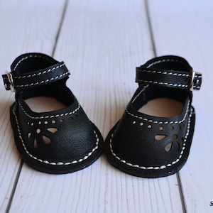 May include: A pair of black leather doll shoes with white stitching and a buckle closure. The shoes have a cut-out design and are made for dolls.